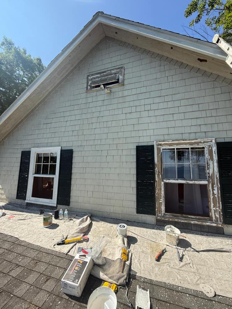 Exterior painting on a shingle-sided gable, one window trim freshly repainted while the next is still to be prepped.