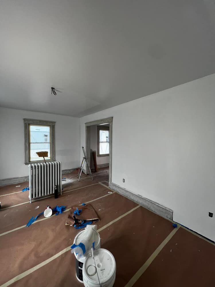 An interior room mid-repaint, fresh coat on the walls and ceiling with the floor papered and the radiator masked.
