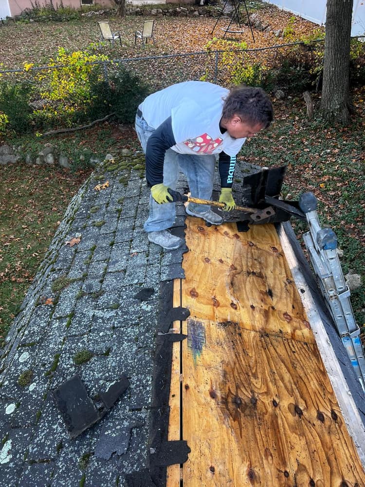 A crew member stripping old moss-covered shingles down to the bare plywood deck.